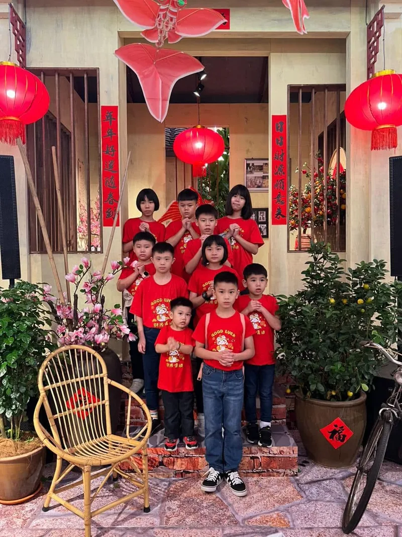 A group of children in matching red 'Good Luck' T-shirts posing under a decorated Chinese New Year gate with lanterns and blossom branches
