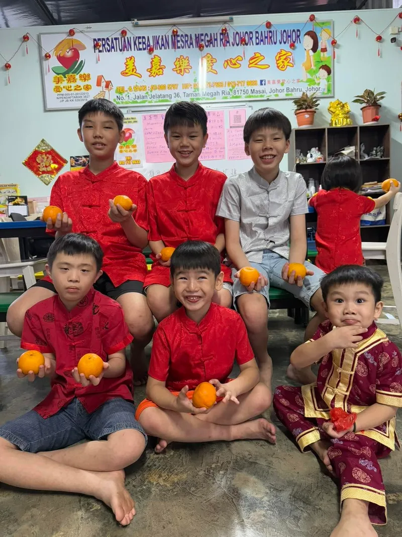 Seven children in red Chinese New Year outfits posing together with mandarin oranges in front of the shelter's noticeboard