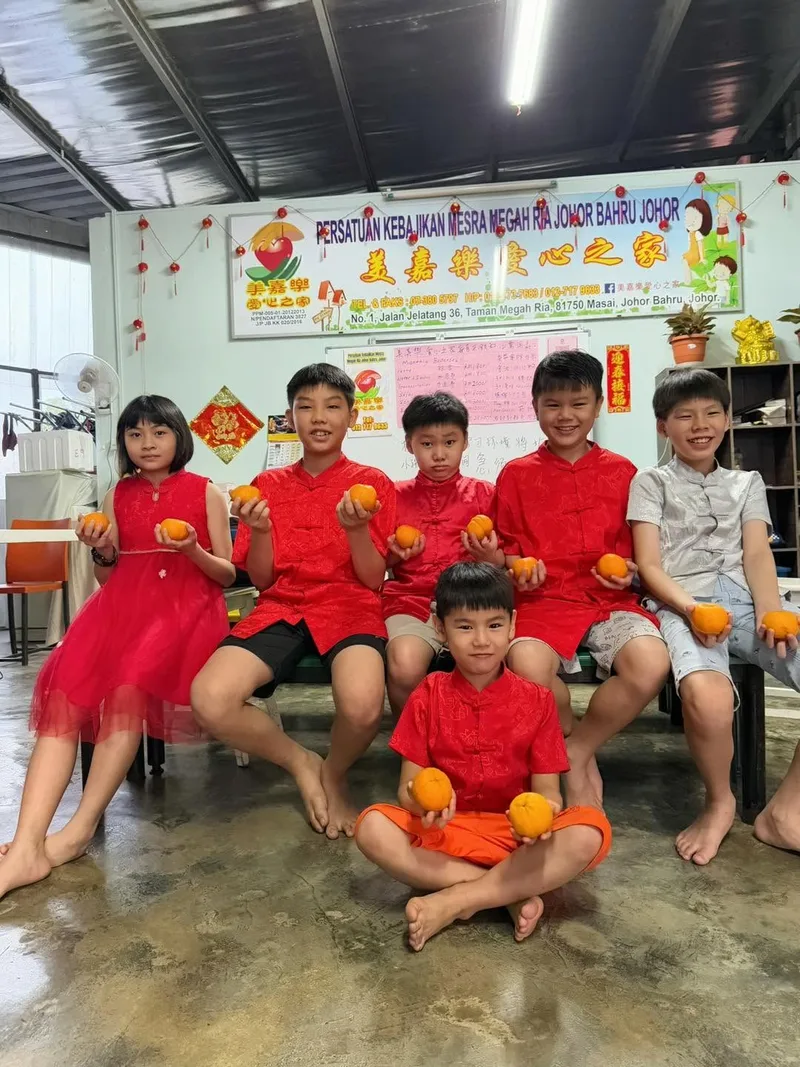 Six children in red Chinese New Year outfits sitting together, each holding mandarin oranges, in front of the shelter's signage