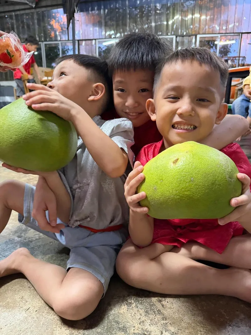Three boys sitting together, two of them holding large pomelos, smiling for the camera